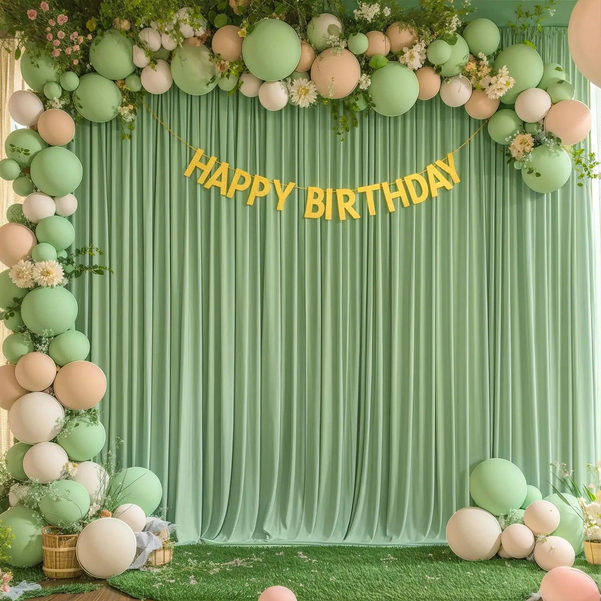 A green draped backdrop decorated with light pink and sage green balloons and greenery for a birthday celebration.