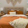 A handwoven boho macrame wall hanging tapestry in cream and beige hangs above a tan upholstered bed.