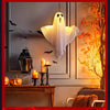 A white ghost light decoration is hanging on a wall in a dimly lit room with autumn foliage visible.