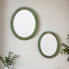 A round, oval wall mirror with a green frame and gold accents hanging on a light gray wall.