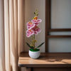 A realistic artificial orchid potted plant with pink flowers sits on a wooden table next to a sheer curtain.