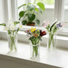 A bouquet of hand-blown glass flowers in various colors displayed in clear mini glass vases on a windowsill.