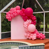 A pink balloon arch decoration set featuring various shades of pink and white balloons in a greenhouse setting.