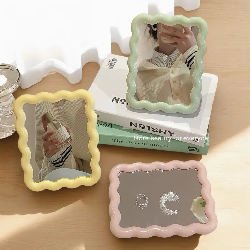 A set of wavy frame makeup mirrors, including a large framed mirror and a smaller pink square mirror, displayed on a wooden s