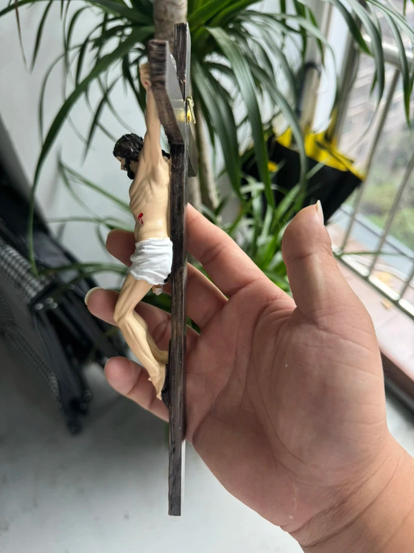 A hand holds a resin sculpture of an Easter cross crucifix wall hanging hanging from a branch amidst green foliage.