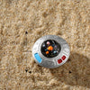Décoration de gâteau d'astronaute miniature avec un disque argenté et des accents colorés posé sur du sable grossier.