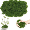A hand holds a clump of bright green artificial moss, with a large mass of similar moss visible above.