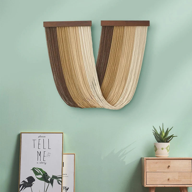 Boho macrame wall tapestry featuring vertical woven fibers in beige and brown tones mounted on a light green wall.