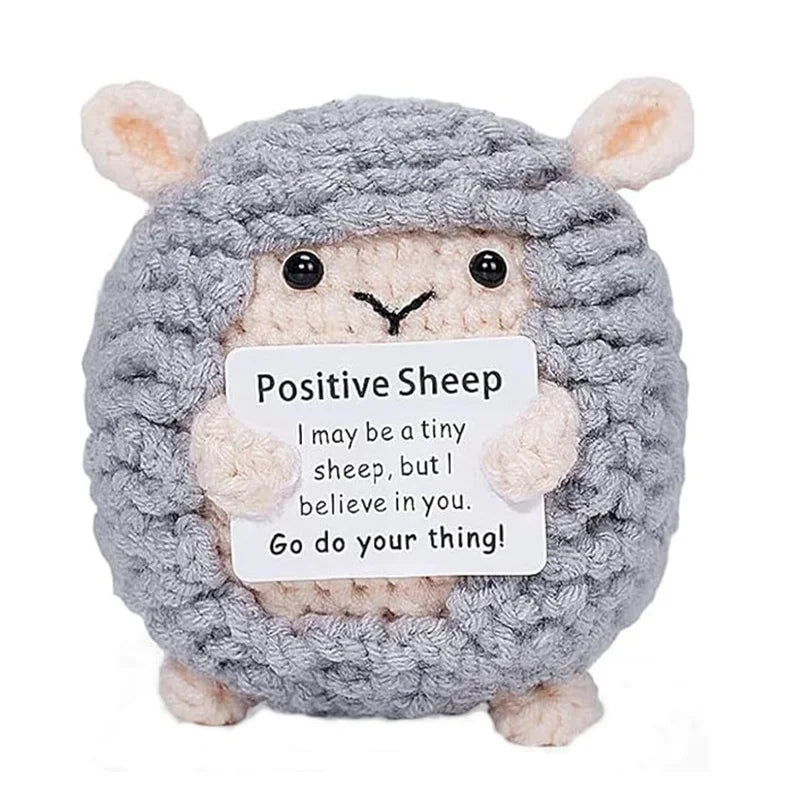 A handmade gray crochet sheep doll with pink ears holding an inspirational card.