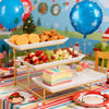 A gold-toned 3-tier serving rack displaying various foods like bread, fried chicken, and fruit on a colorful striped tableclo