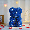 Blauer künstlicher Rosen-Teddybär, umhüllt von blauen Rosen, auf weißer Fläche mit sanftem Licht.