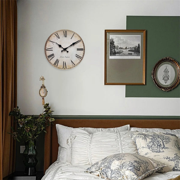 A round wooden wall clock with Roman numerals hangs on a white wall above a bed.