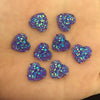 Eine Sammlung von 12mm herzförmigen Strasssteinen mit irisierendem blau-violettem Glitzer, verstreut auf einer Hand.