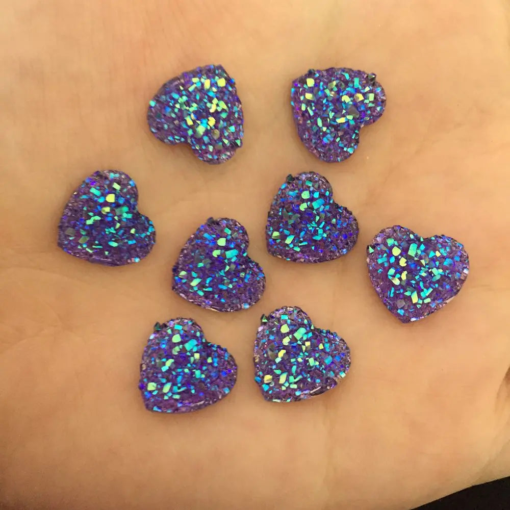 Eine Sammlung von 12mm herzförmigen Strasssteinen mit irisierendem blau-violettem Glitzer, verstreut auf einer Hand.