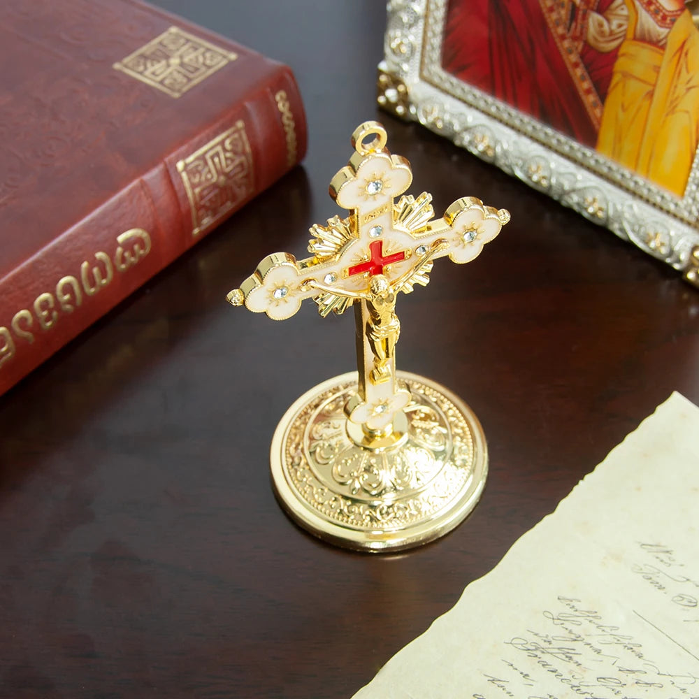 A golden metal Jesus cross ornament with a decorative base, resting on a dark wooden surface next to a book and framed artwor
