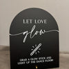 A black acrylic sign with white script that reads Let Love Glow and instructs to grab a glow stick and light up the dance flo