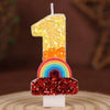 A single golden number one candle with a rainbow sequined base on a wooden surface.