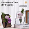 Clear acrylic photo frame vase with greenery and flowers on a wooden surface.