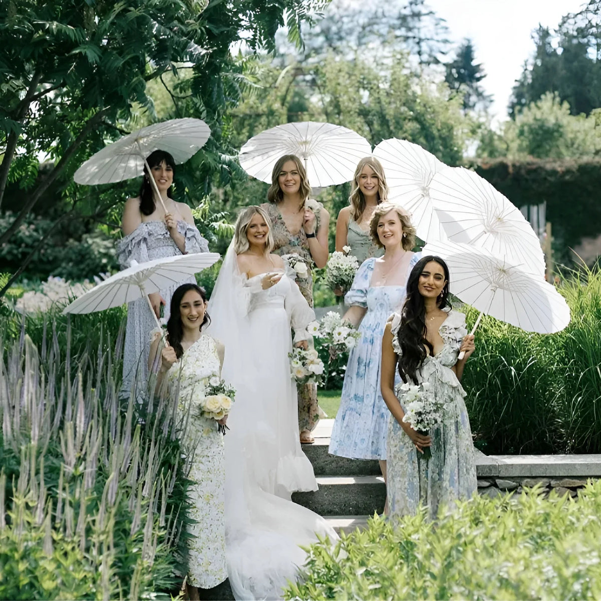 A bride and bridesmaid hold white oil paper umbrellas in a garden setting.