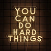 A glowing neon sign spelling out YOU CAN DO HARD THINGS against a dark grid background.