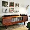 A wooden cabinet with a glass top and a section for vinyl records is displayed against a white wall with framed art.