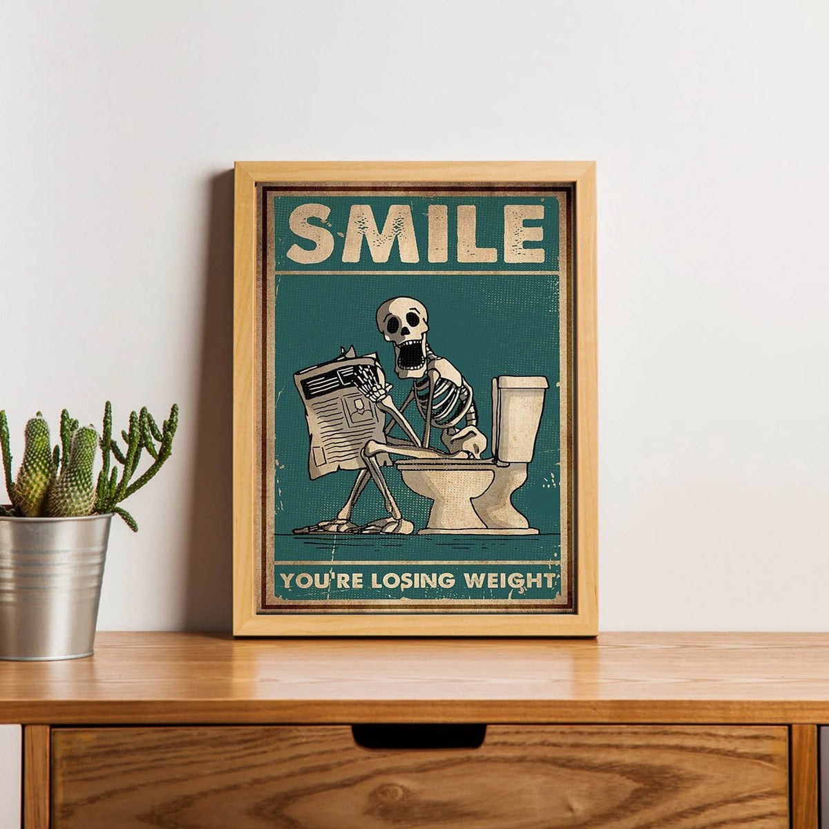 A framed canvas print featuring a vintage illustration of a skeleton squatting on a toilet with the text You're losing weight