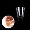 A hand holds a small glass of amber liquid next to a clear plastic shot glass.