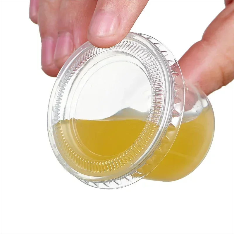 A hand is holding an open clear plastic sauce cup with a lid, showing a yellowish liquid inside.