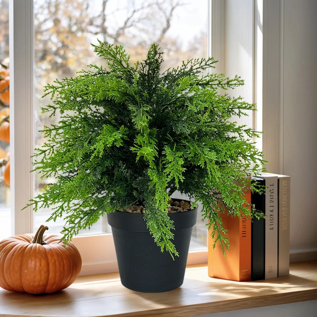 A potted artificial cypress topiary with bright green foliage sits on a windowsill next to a pumpkin and a stack of books.