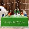 A green cake decorated with a soccer field theme featuring figurines, candles shaped like numbers, and the words Happy Birthd