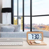 A solid wood digital alarm clock displaying 7:30, with a calendar and temperature display, resting on a desk next to a laptop