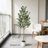 A tall artificial olive tree in a white pot standing indoors next to a window and wooden furniture.