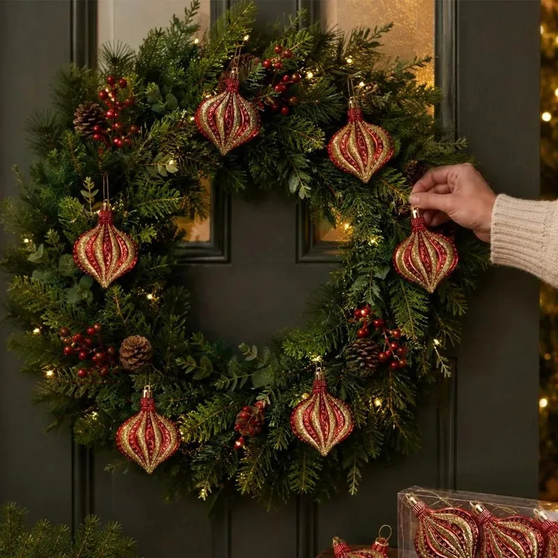 A festive wreath of green pine branches decorated with red ornaments and pinecones, being held by a hand.