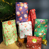 A collection of Christmas kraft paper gift bags with seal stickers in various colors and patterns displayed next to a pine br