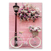 A pink bicycle with a basket of flowers is displayed against a brick background.
