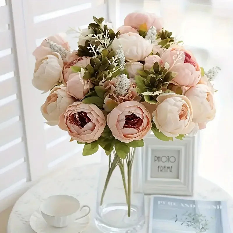 A bouquet of pink and cream peony artificial flowers with green foliage displayed in a glass vase.