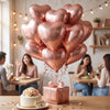 A bouquet of pink heart-shaped foil balloons is displayed on a wooden table with a cake and flowers.