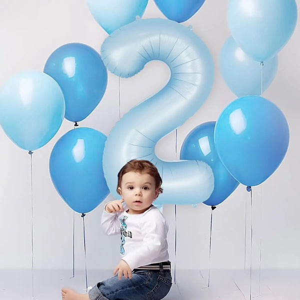 A toddler is sitting in front of a large light blue foil number balloon shaped like the number 2.