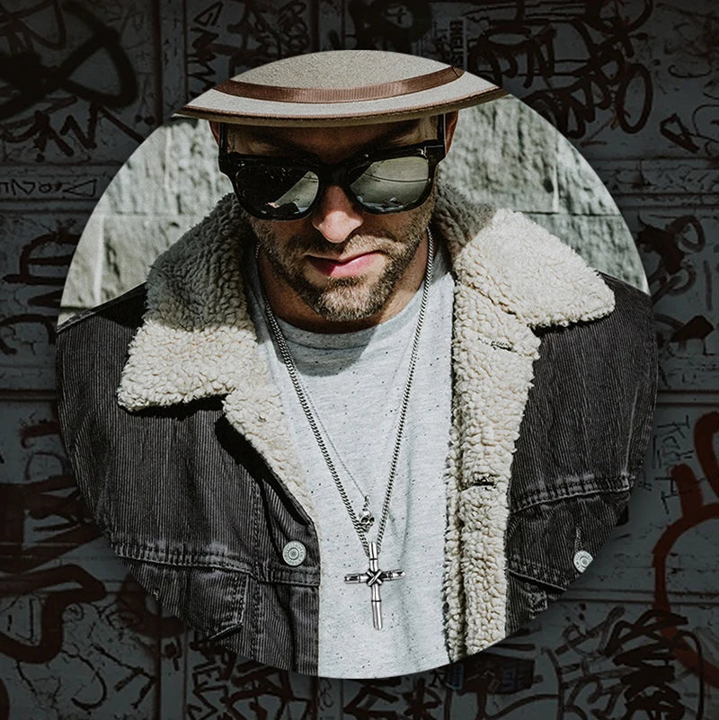 A man wearing sunglasses and a hat is shown in a close-up portrait, featuring a stainless steel cross necklace pendant.