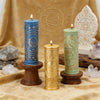 Three pillar candles featuring celestial and mystical symbols in blue, gold, and green on a beige fabric.