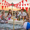 A group of people enjoying an outdoor party under a red triangle bunting flag canopy at sunset.