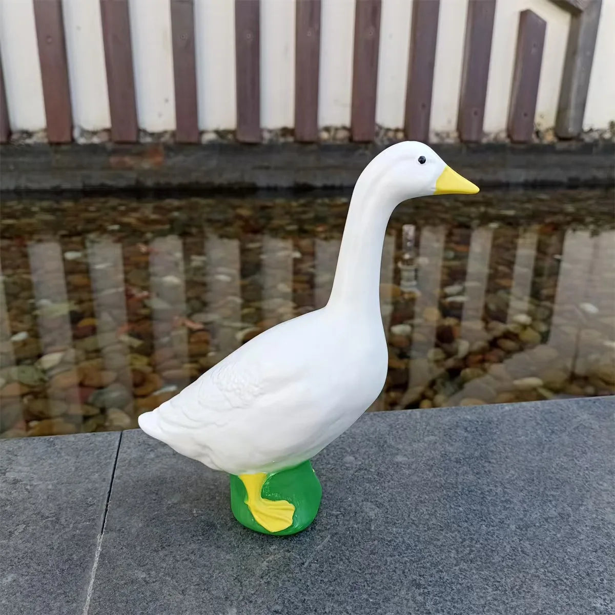 A white garden statue of a goose with a yellow beak and green feet is placed on a dark surface.