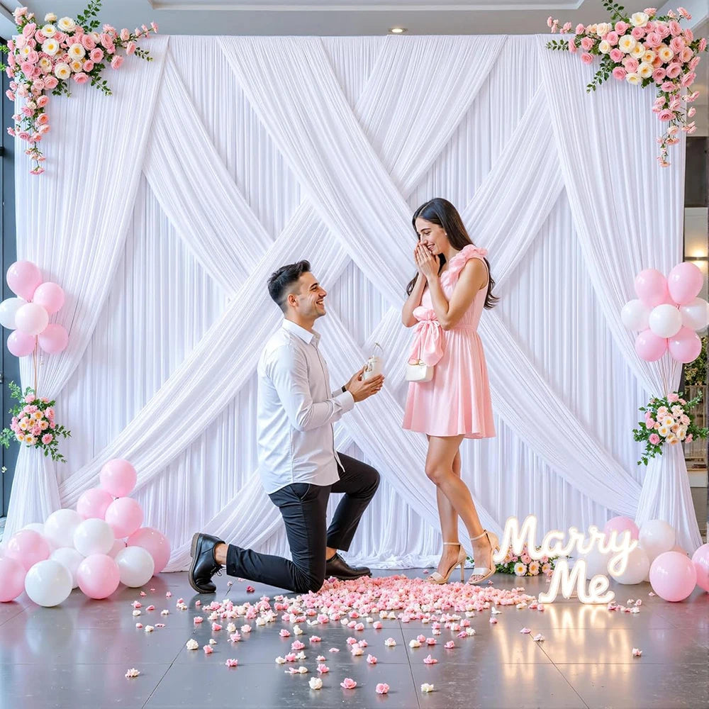 A bride and groom pose in front of a white draped fabric backdrop decorated with pink and white flowers and scattered petals.