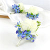 A set of six dusty blue silk rose boutonnières and white roses arranged on a white cloth.