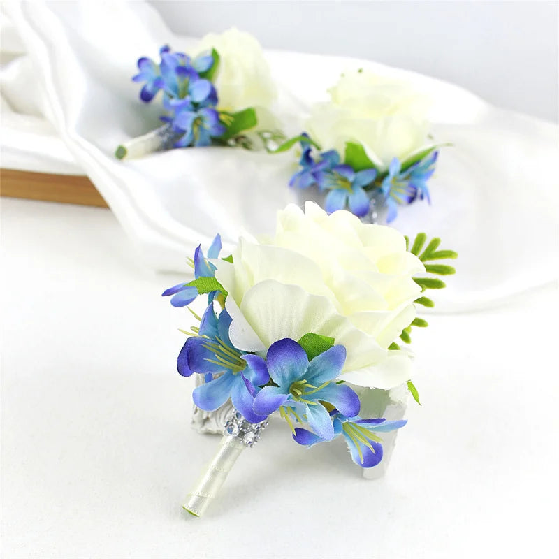 A set of six dusty blue silk rose boutonnières and white roses arranged on a white cloth.