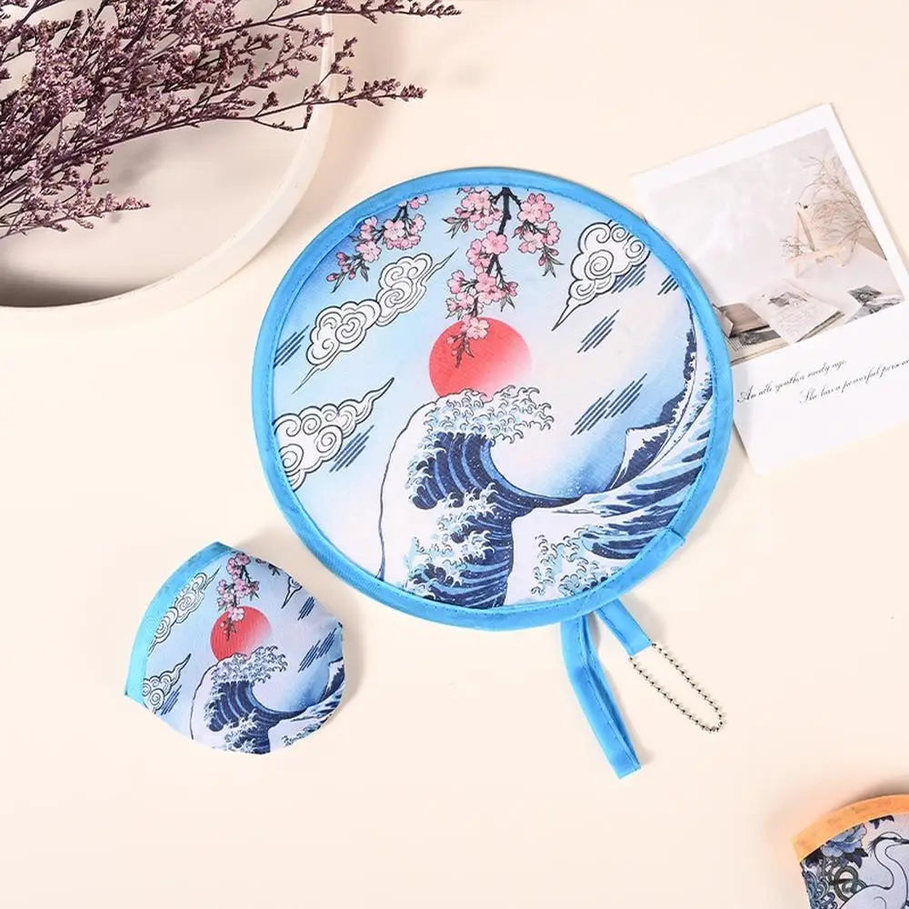 A blue portable foldable cooling handheld fan with a wave design and pink cherry blossoms is shown on a light background.