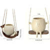 A beige ceramic mug ornament shaped like a smiling face, hanging from a string on a wooden base.