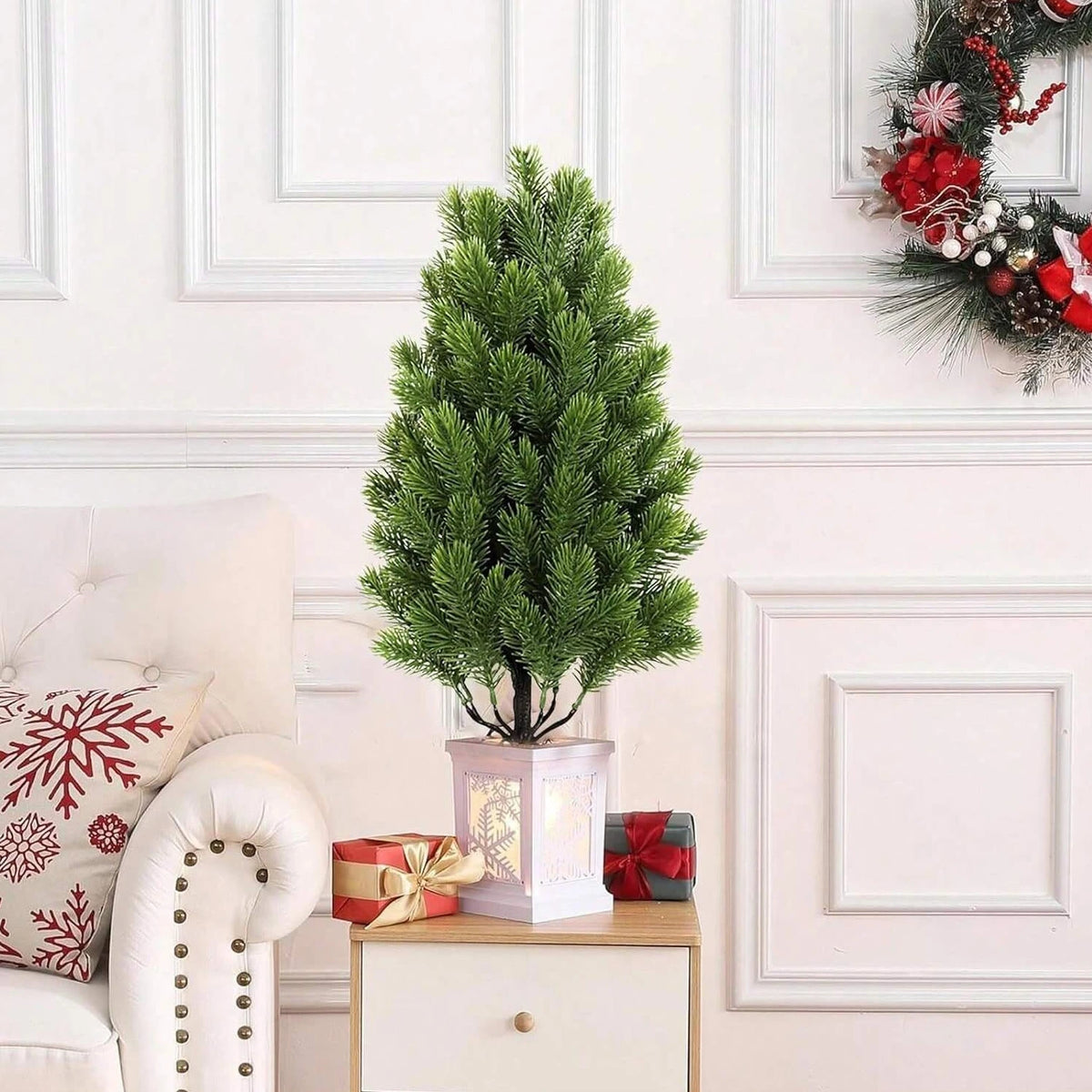 A tall artificial pine tree in a white planter decorated with holiday accents, set against a white wall.