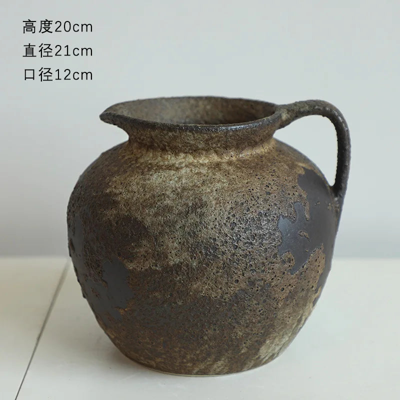 A dark, textured ceramic pitcher or vase with a handle and spout is shown against a light background.