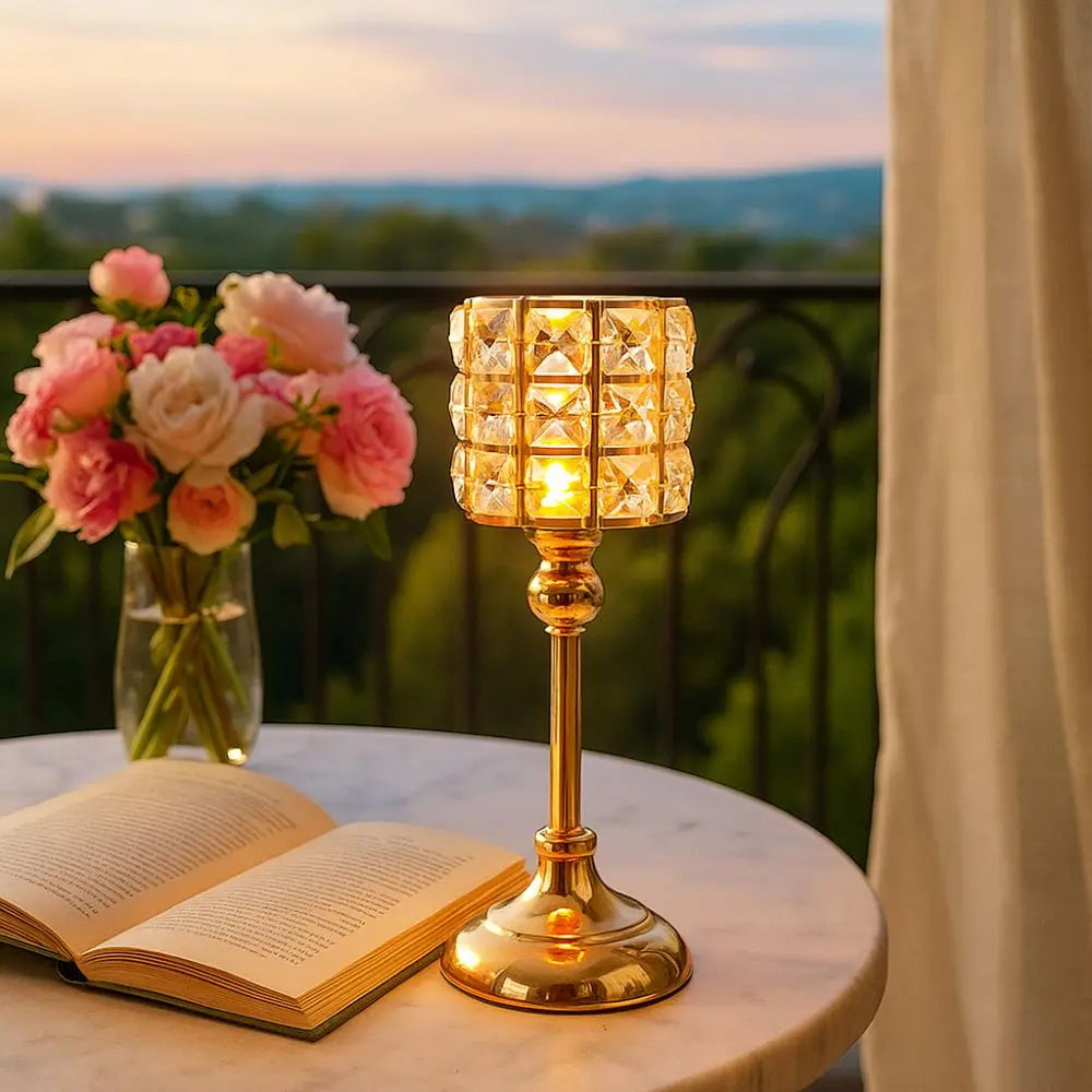 Portavelas de cristal dorado con vela encendida sobre una mesa de mármol junto a un libro abierto y un atardecer.